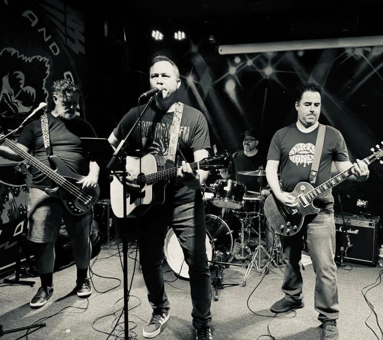 The Rewind Theory playing live: Kevin on bass, Chris singing with an acoustic guitar, Roy on drums and Justin on lead guitar