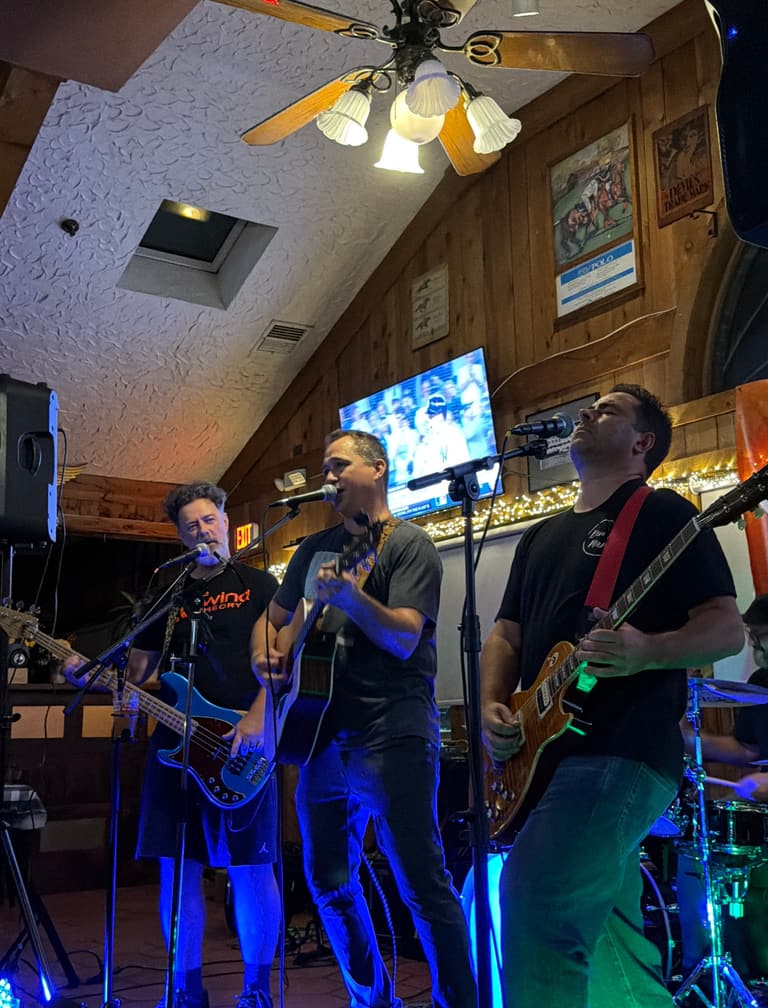 The Rewind Theory live in a wood-paneled room: Kevin on bass, Chris singing with an acoustic guitar and Justin on lead guitar